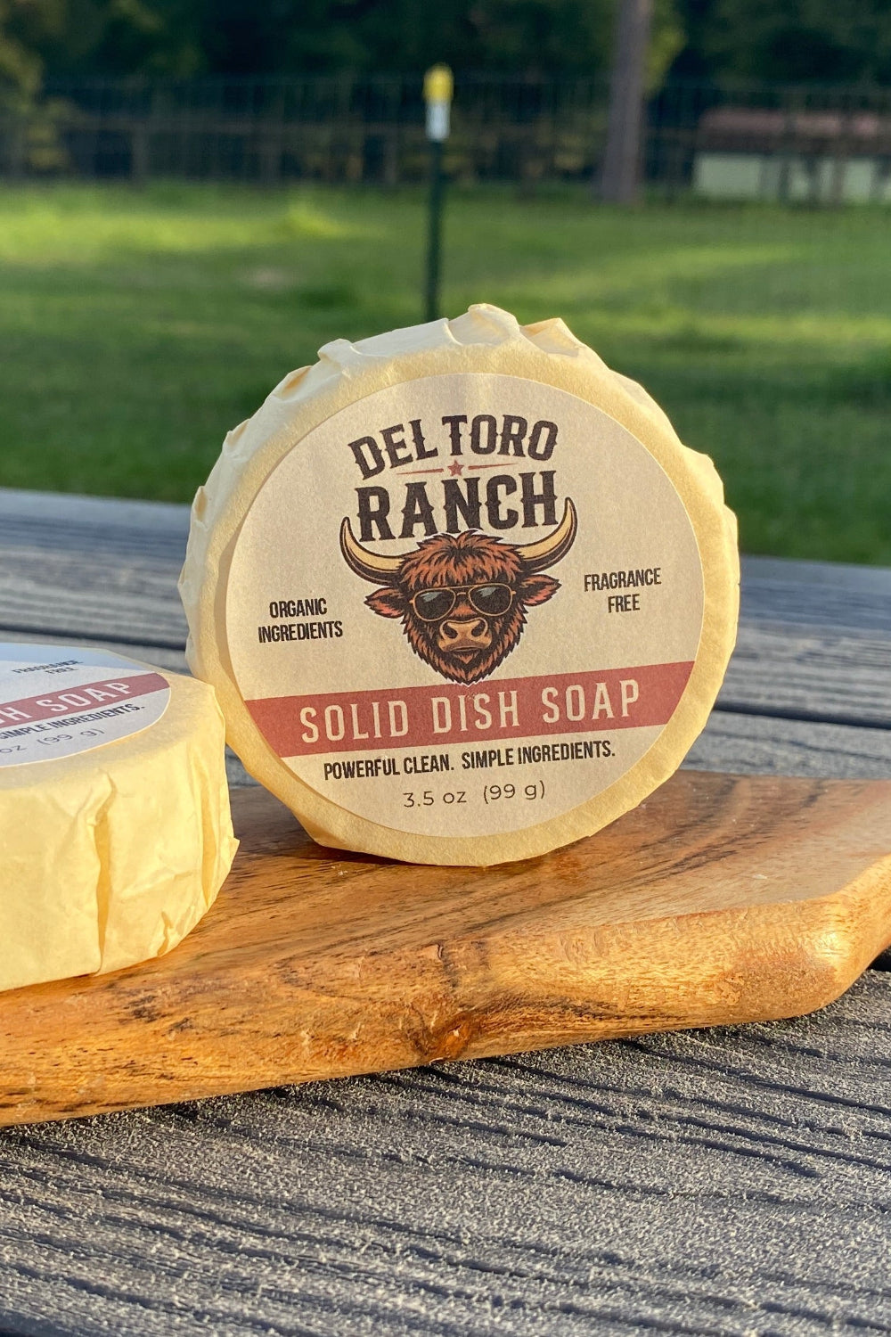 Two pucks of Del Toro Ranch solid dish soap displayed on a wooden cutting board on a gray picnic table, with a blurred green grassy field and trees in the background.
