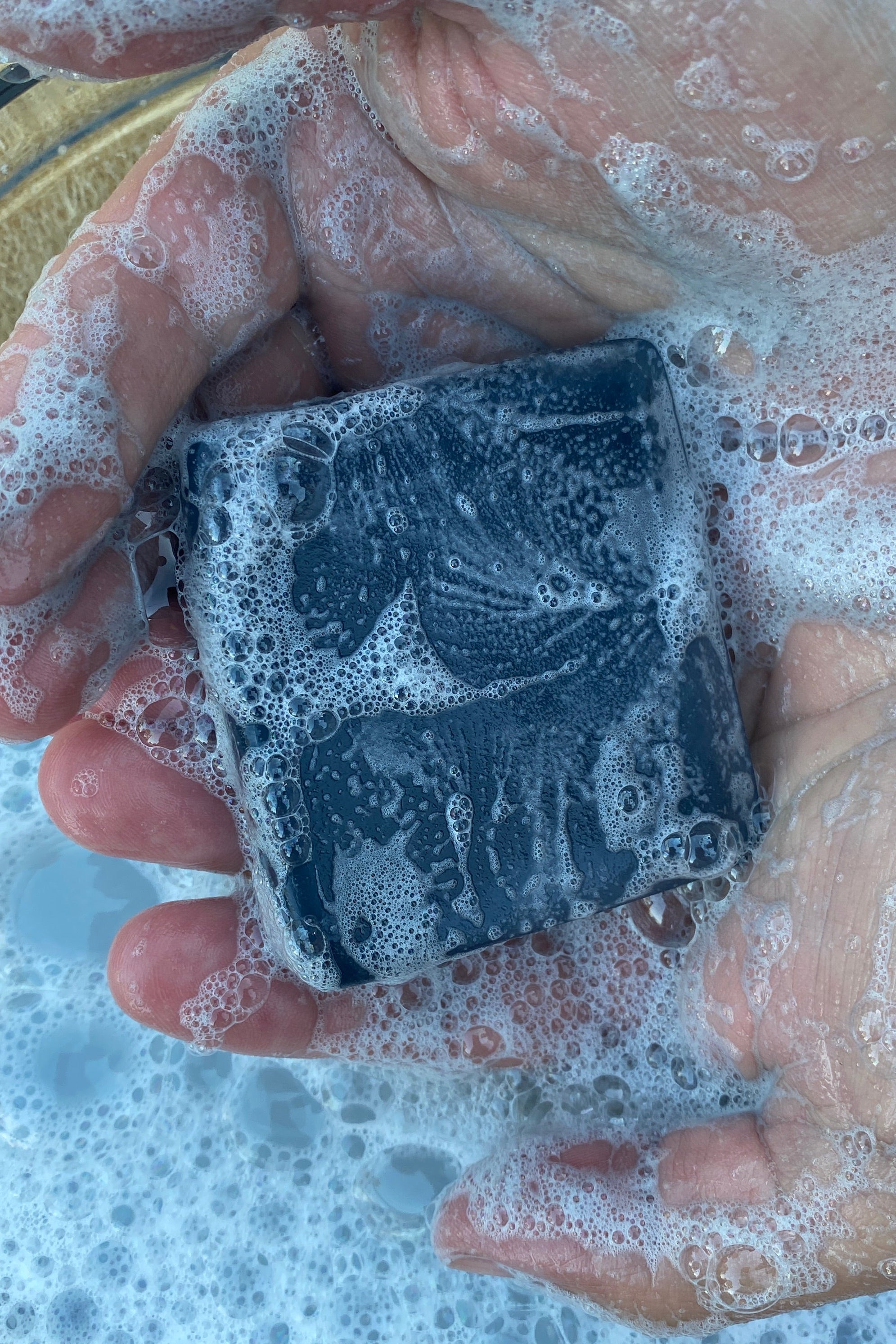 A close-up of a person's hands covered in sudsy bubbles, holding a dark charcoal-colored square bar of the shop bar soap over a bowl of soapy water.