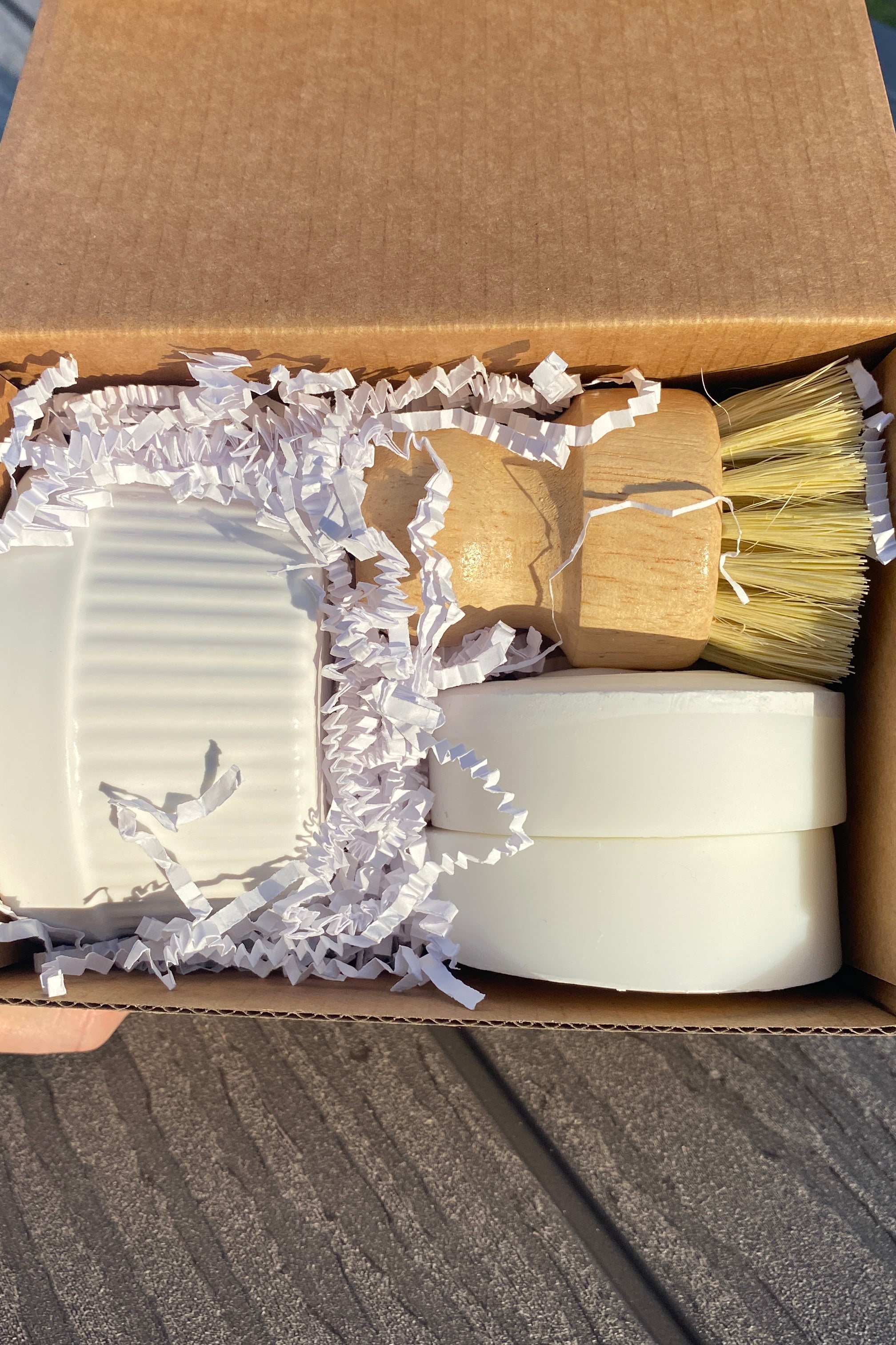 A hand holds an open cardboard gift box containing a white ceramic ramekin with  solid dish soap, two round refills, and one bamboo dish brushb. all surrounded by white paper shred, set on a gray wooden deck.