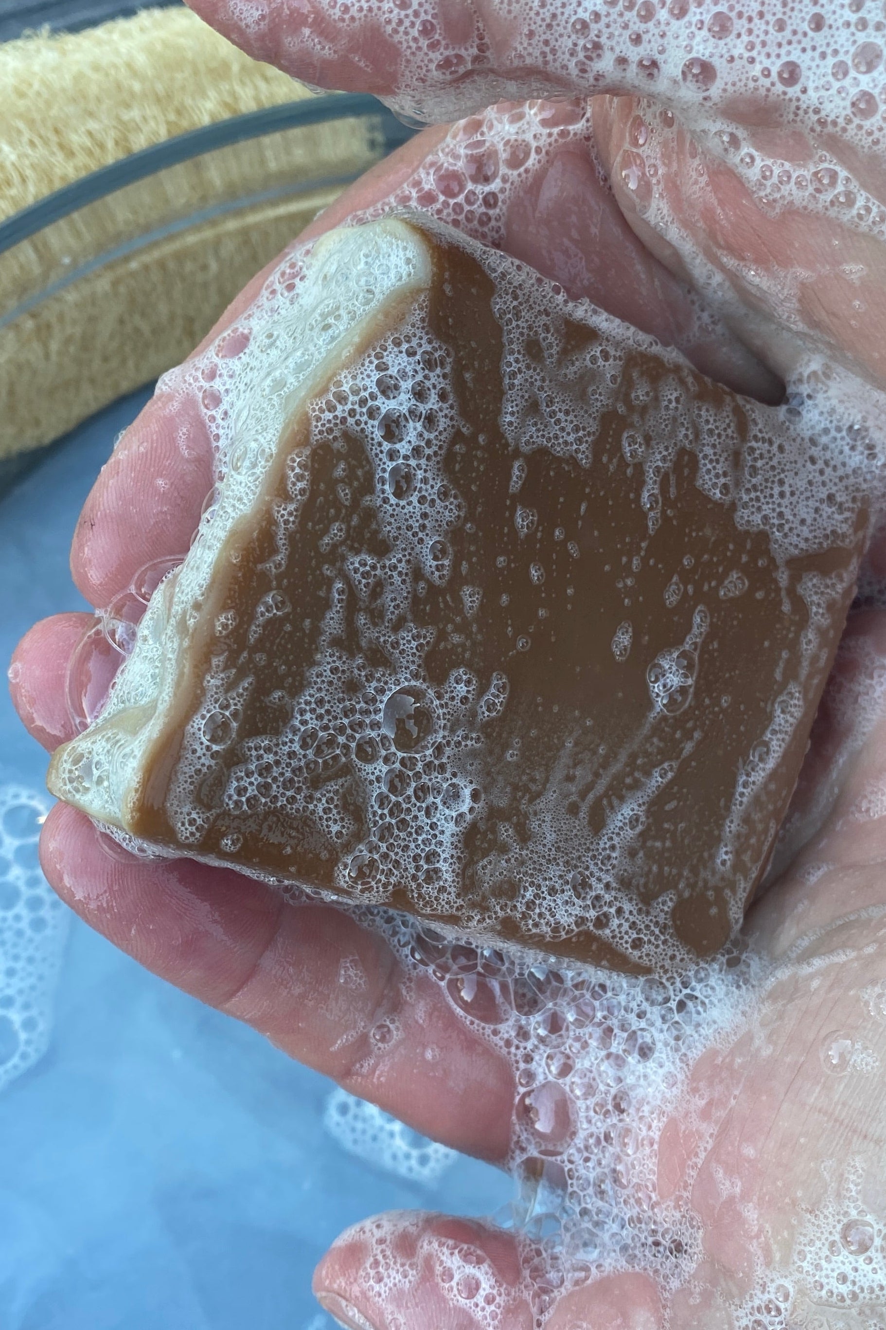 A hand holds a rectangular bar of brown signature cacao soap covered in lather and foam, with a glass bowl of soapy water and a yellow scrubber visible in the background.