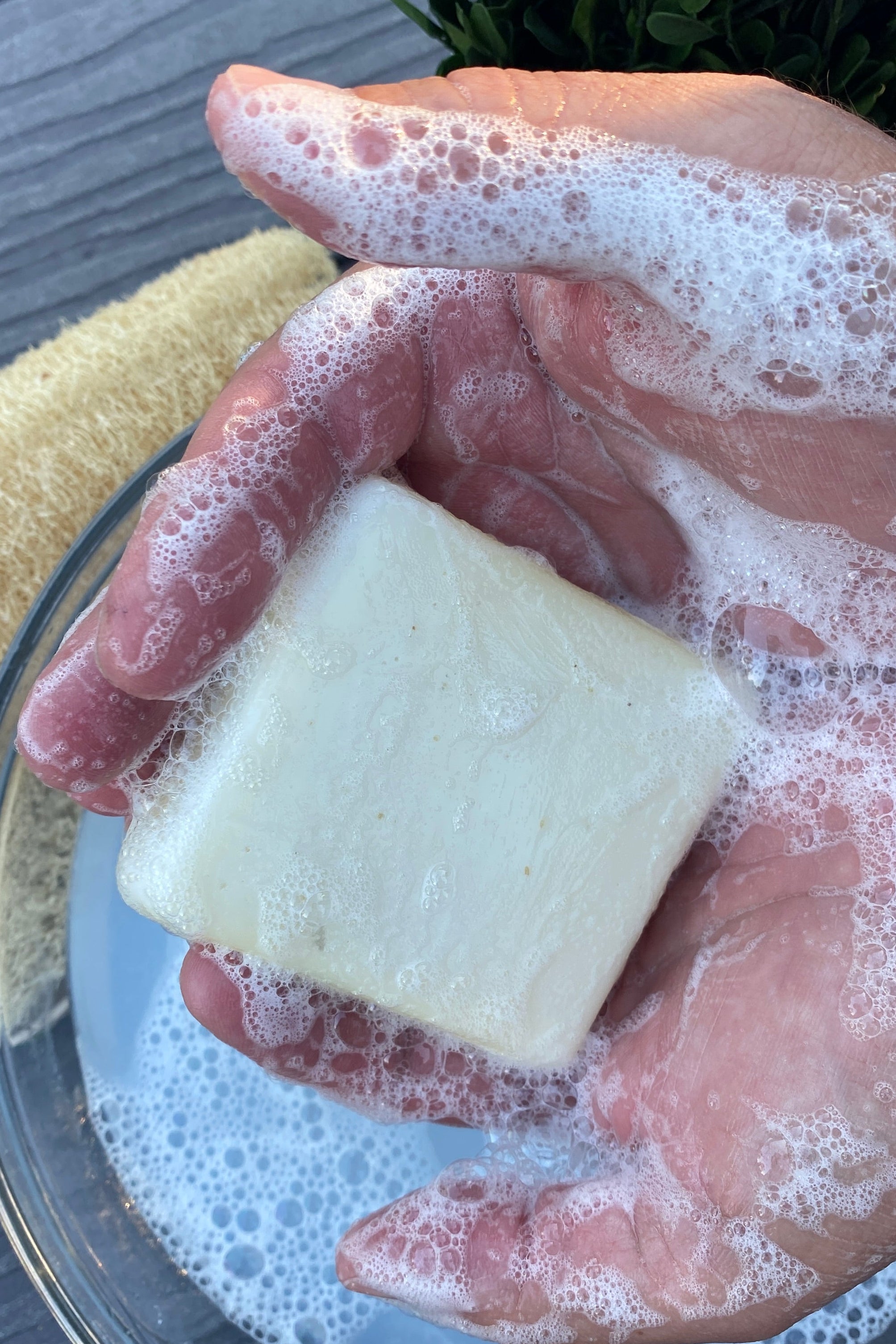 A hand holds a rectangular bar of pale cream colored tallow original soap covered in white lather foam over a glass bowl, with a natural sponge and greenery in the background.